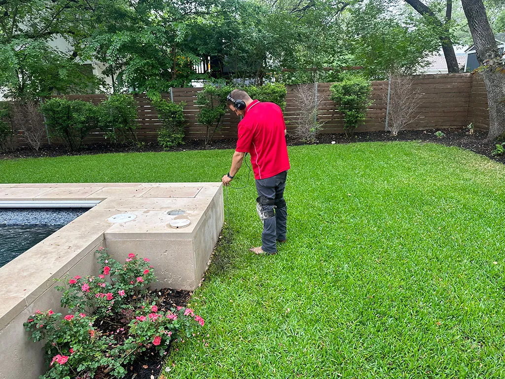 Person in a red shirt using a tool on the edge of a pool in a green backyard with a wooden fence and bushes.