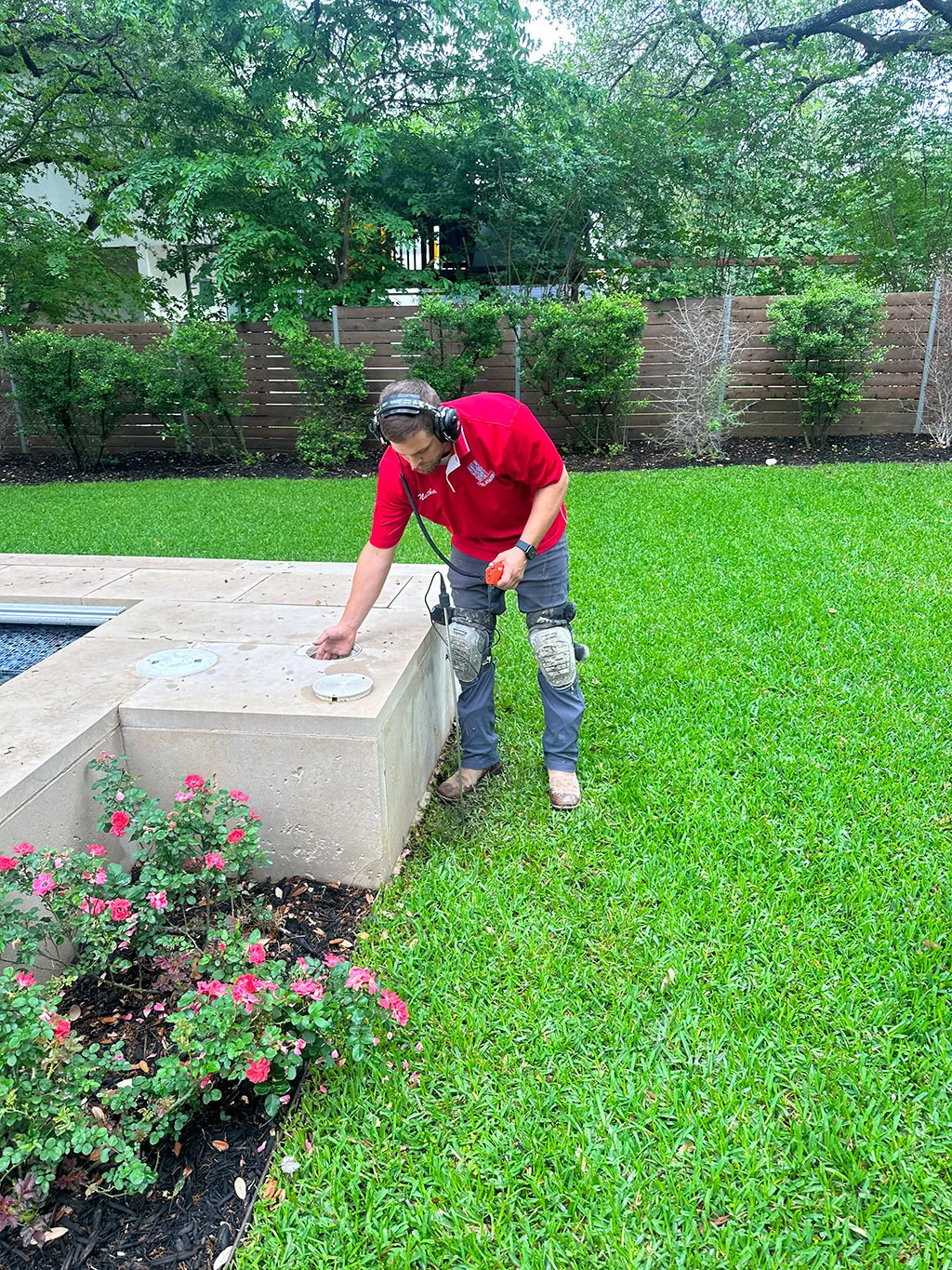 Technician wearing headphones and red shirt inspecting an outdoor irrigation system near a garden and green lawn.