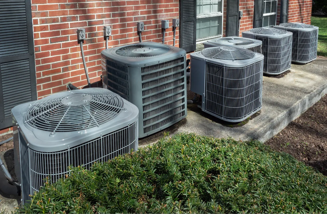 Five outdoor air conditioning units lined up along a brick wall outside a house.