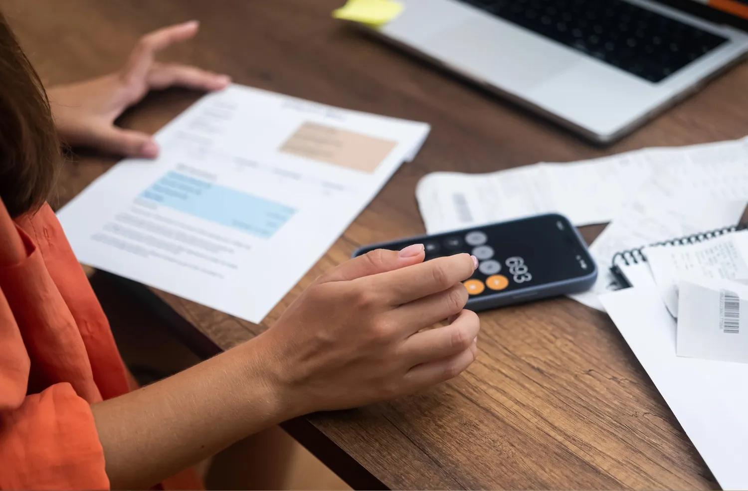 Person calculating finances using a smartphone calculator app, holding a document with highlighted sections, with a laptop and receipts on a wooden desk.