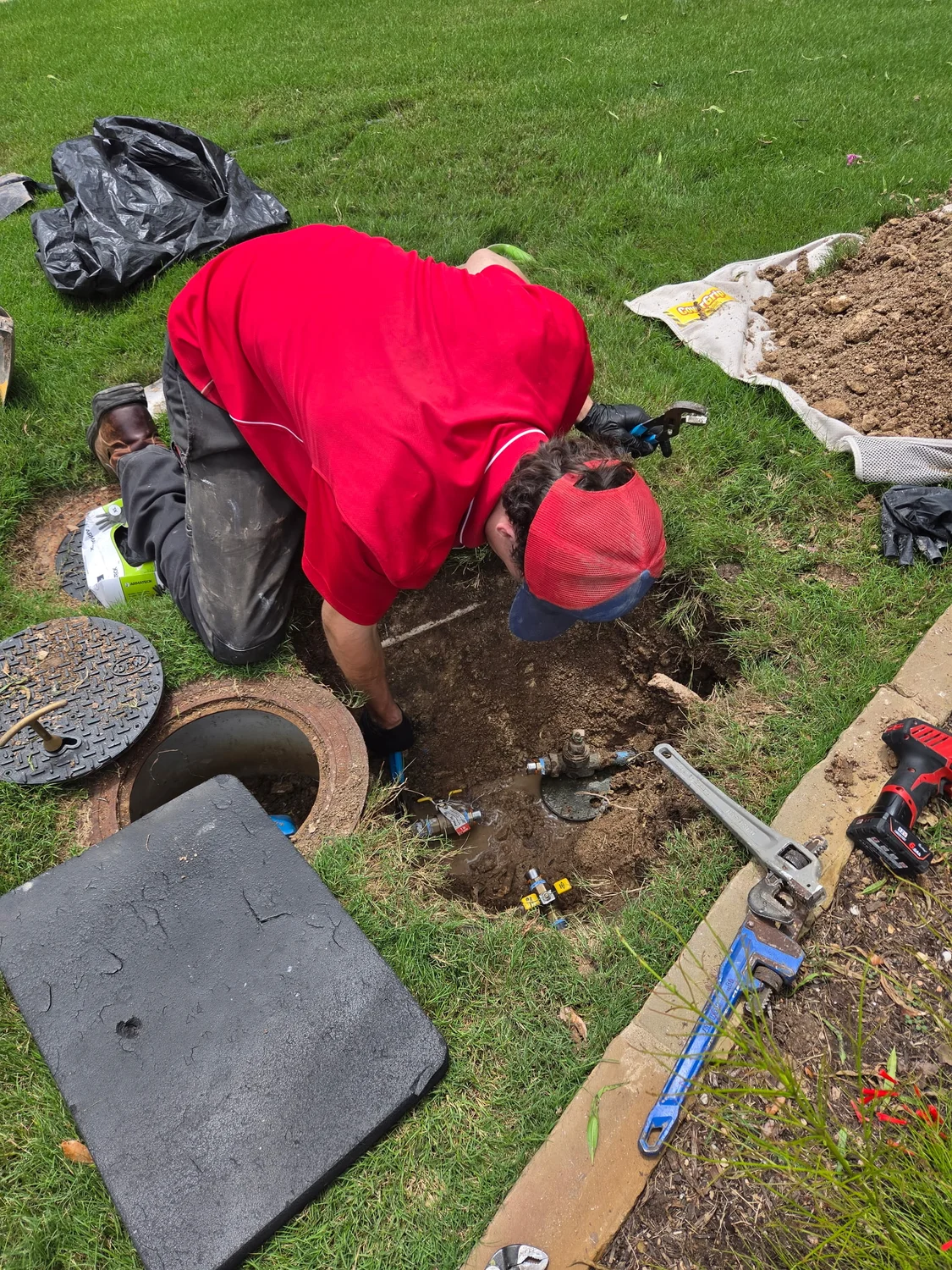 Worker in red shirt repairing underground water pipes with tools on a grassy lawn.