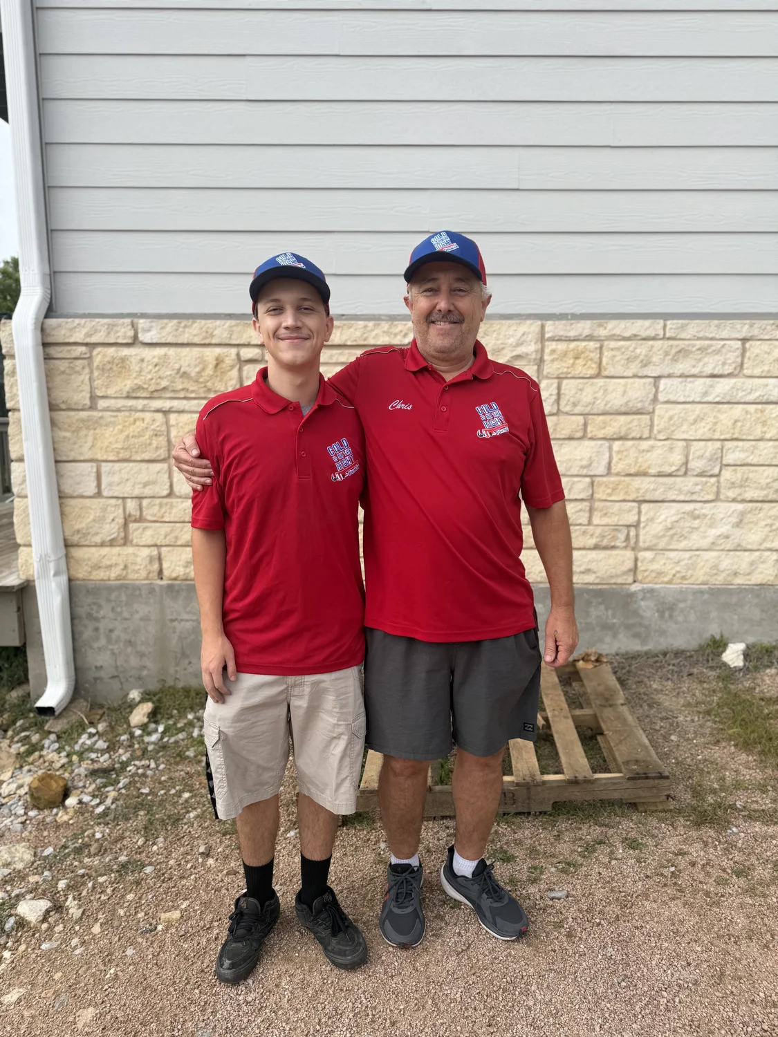 Father and son smiling in matching red shirts and hats, standing outside near a building wall.