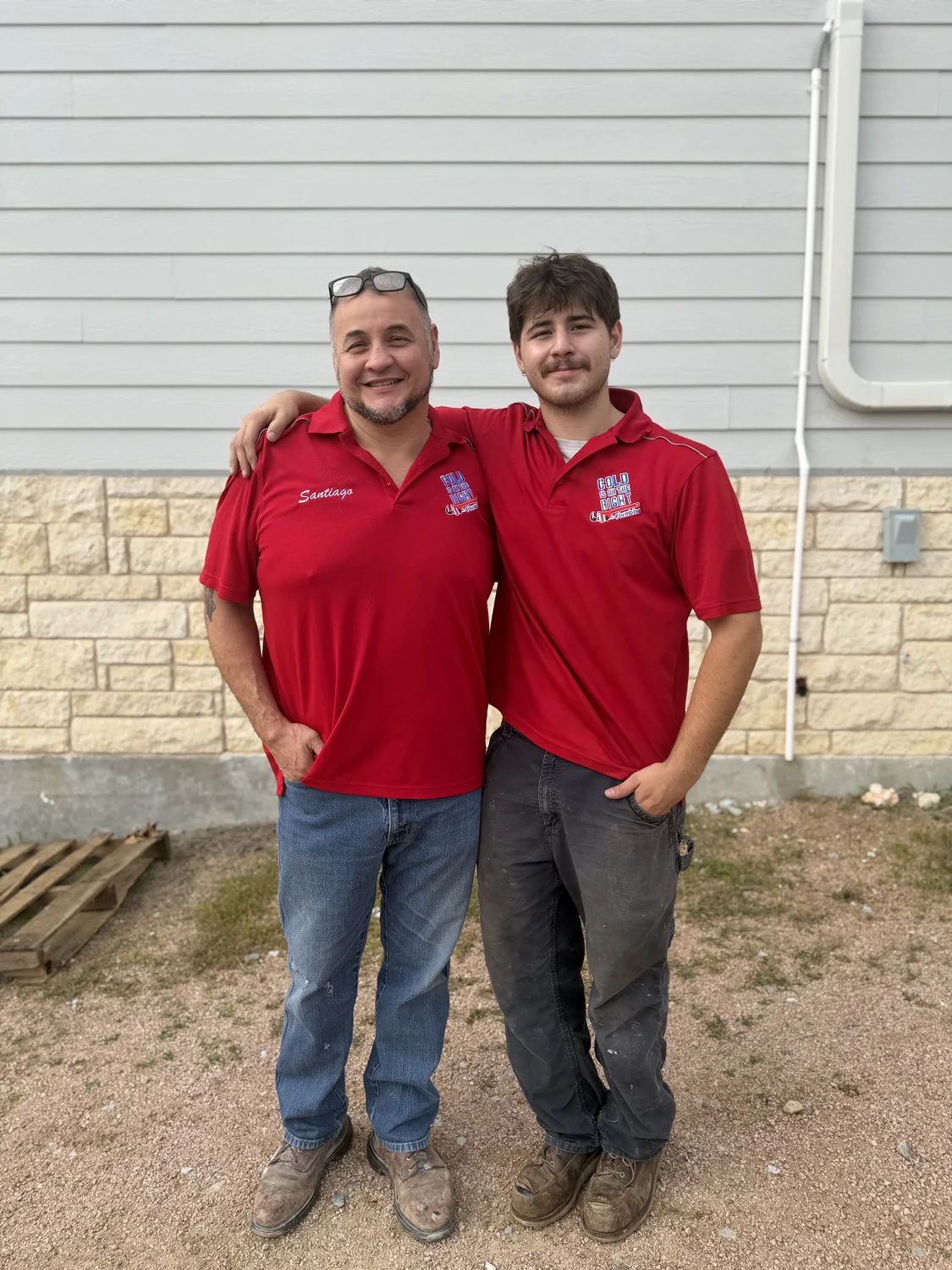 Two men in red work shirts with arms around each other standing outside a building.
