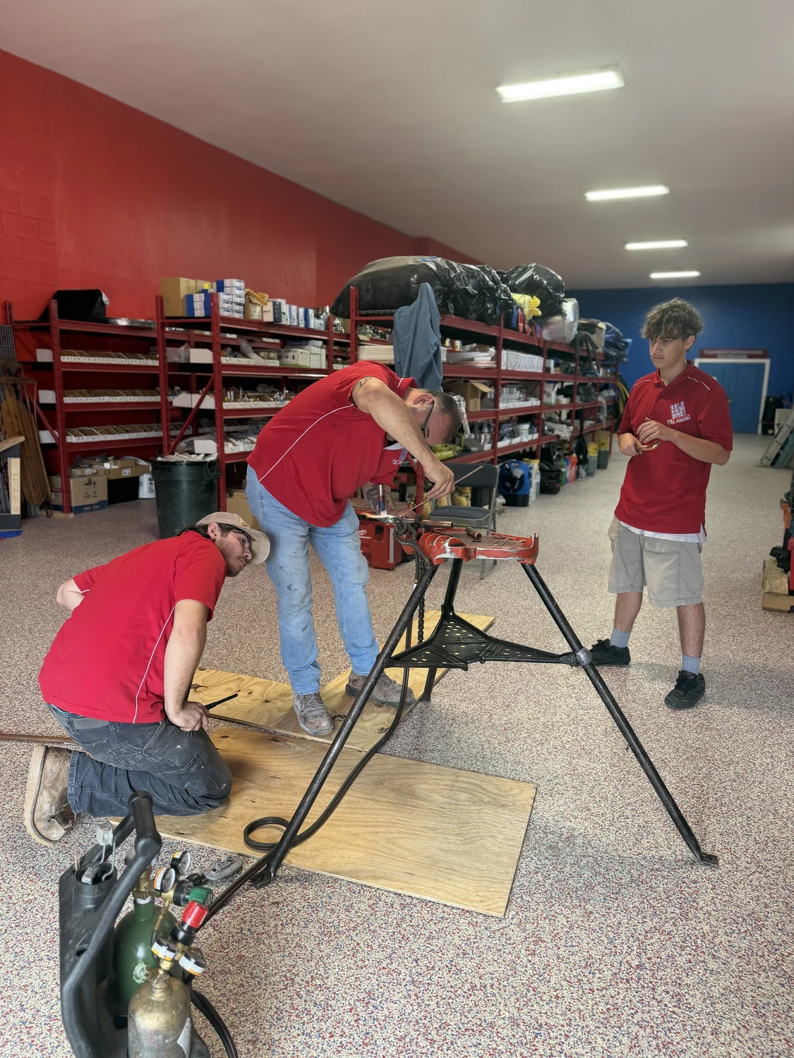 Three men in red shirts working on pipe fitting in a workshop with shelves of supplies in the background.