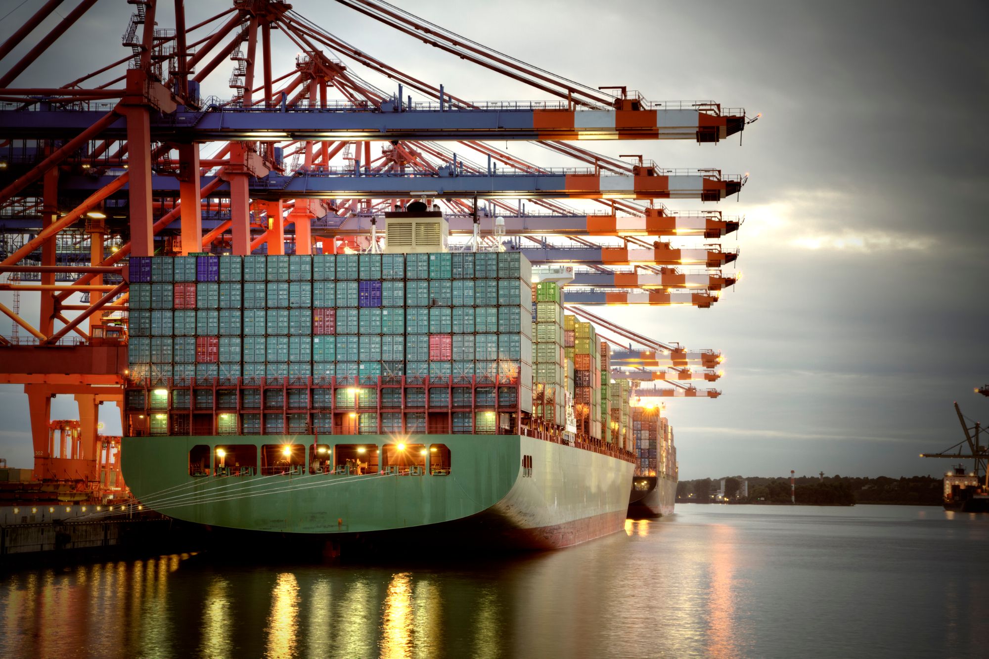 Containerschiff unter Containerbrücken im Hafen bei Dämmerung.