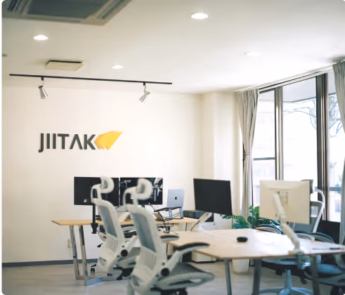 Photo of a jiitak japan office with white chairs, desks, computers, large windows, and the word "Jiitak" on the wall.