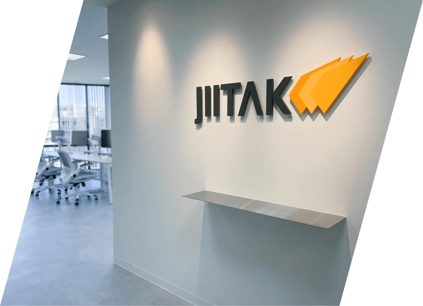 白い壁にロゴがあるJIITAK福岡本社。背景にはモダンなワークスペースが見える。