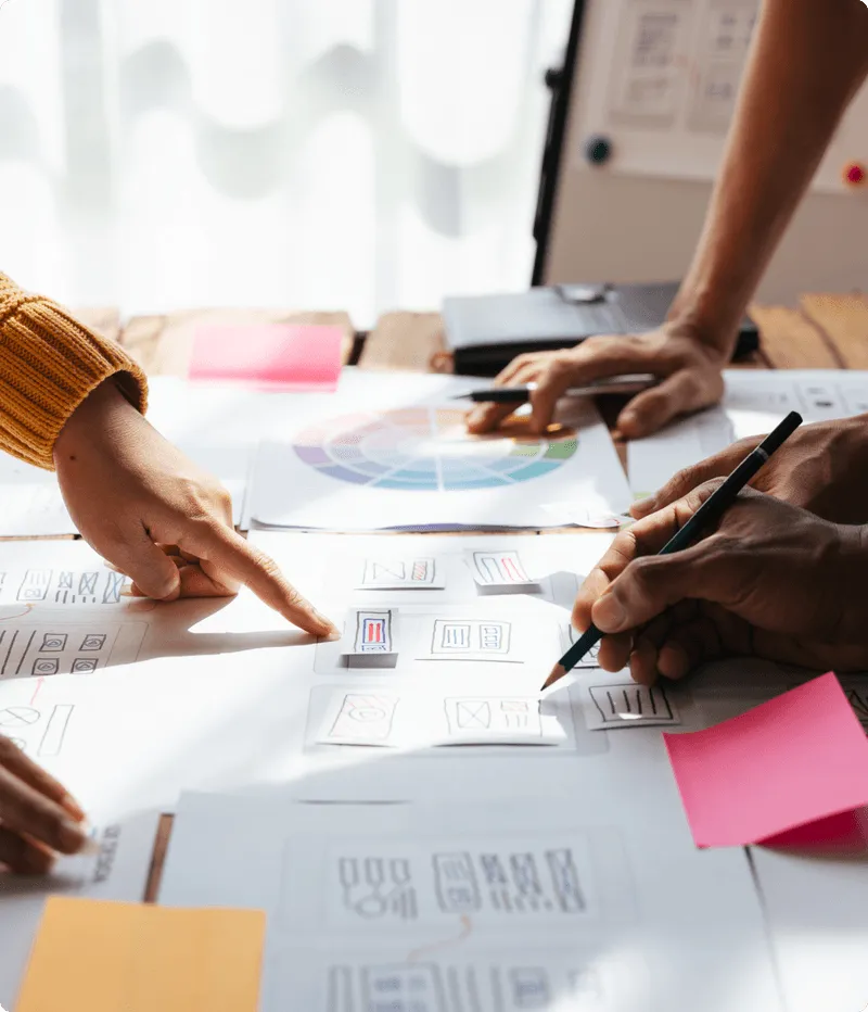 デスクで印刷されたワイヤーフレームスケッチやチャートを確認し、サービス画面のプロトタイプ作成について議論する複数の人々。