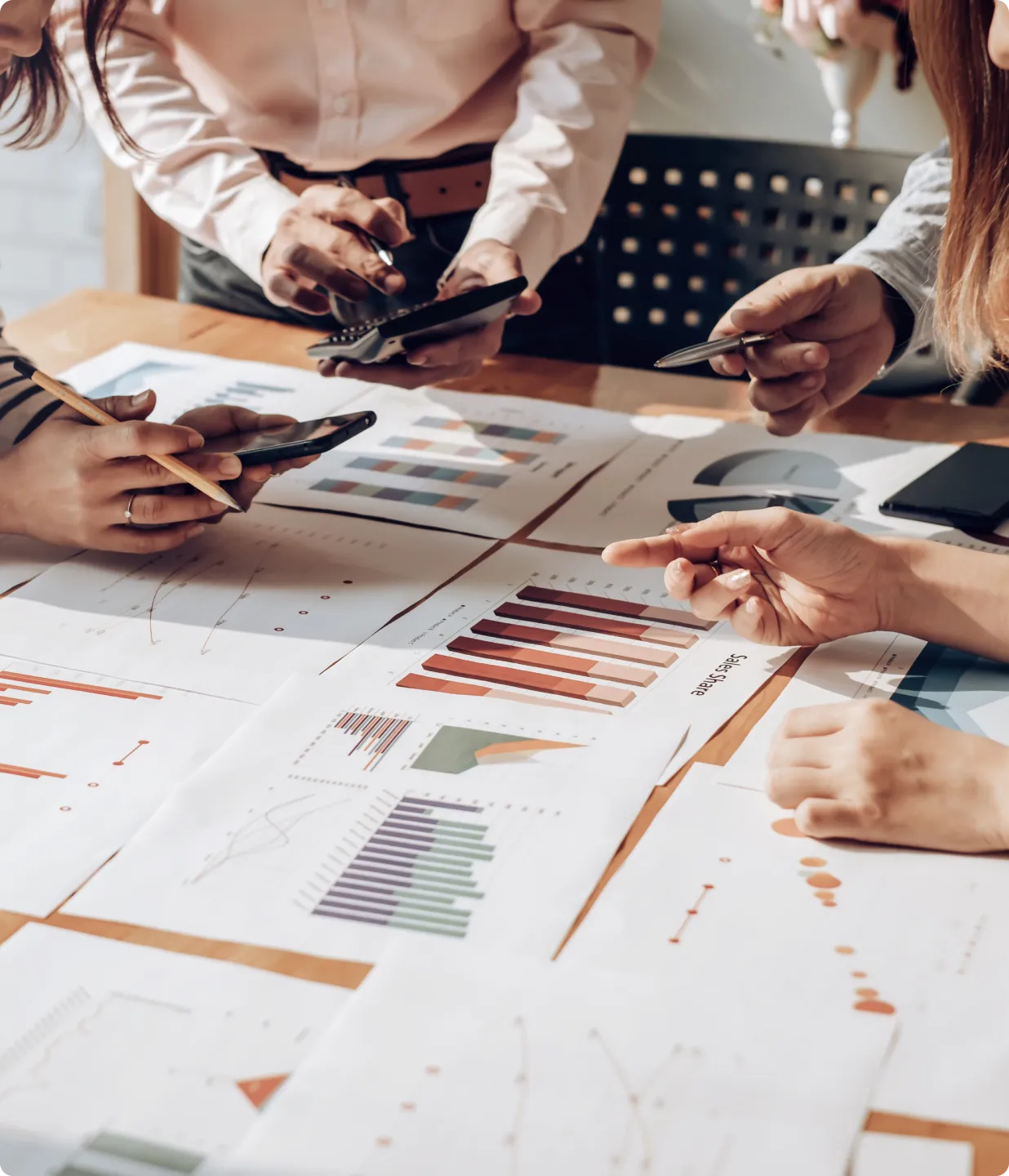 デスクに置かれた印刷されたチャートやグラフを分析し、ペンやスマートフォンを持ちながらフィードバックと評価戦略について議論する複数の人々。