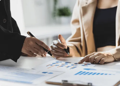 印刷されたチャートやグラフを分析し、ペンでグラフを指しながら実現可能性について議論する2人。