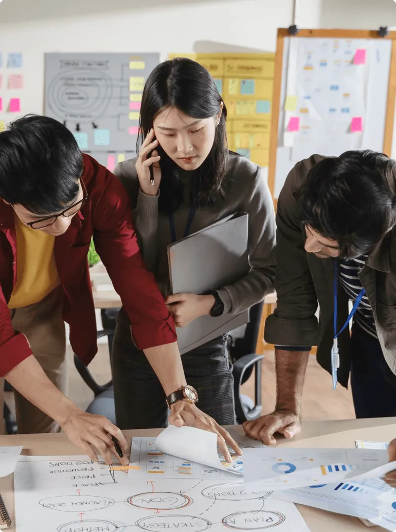 オフィスでホワイトボードを使って協力しながら議論している3人のビジネスパーソン。1人は電話で話しながら資料を持ち、他の2人はホワイトボードに書き込みながら意見を交わしています。