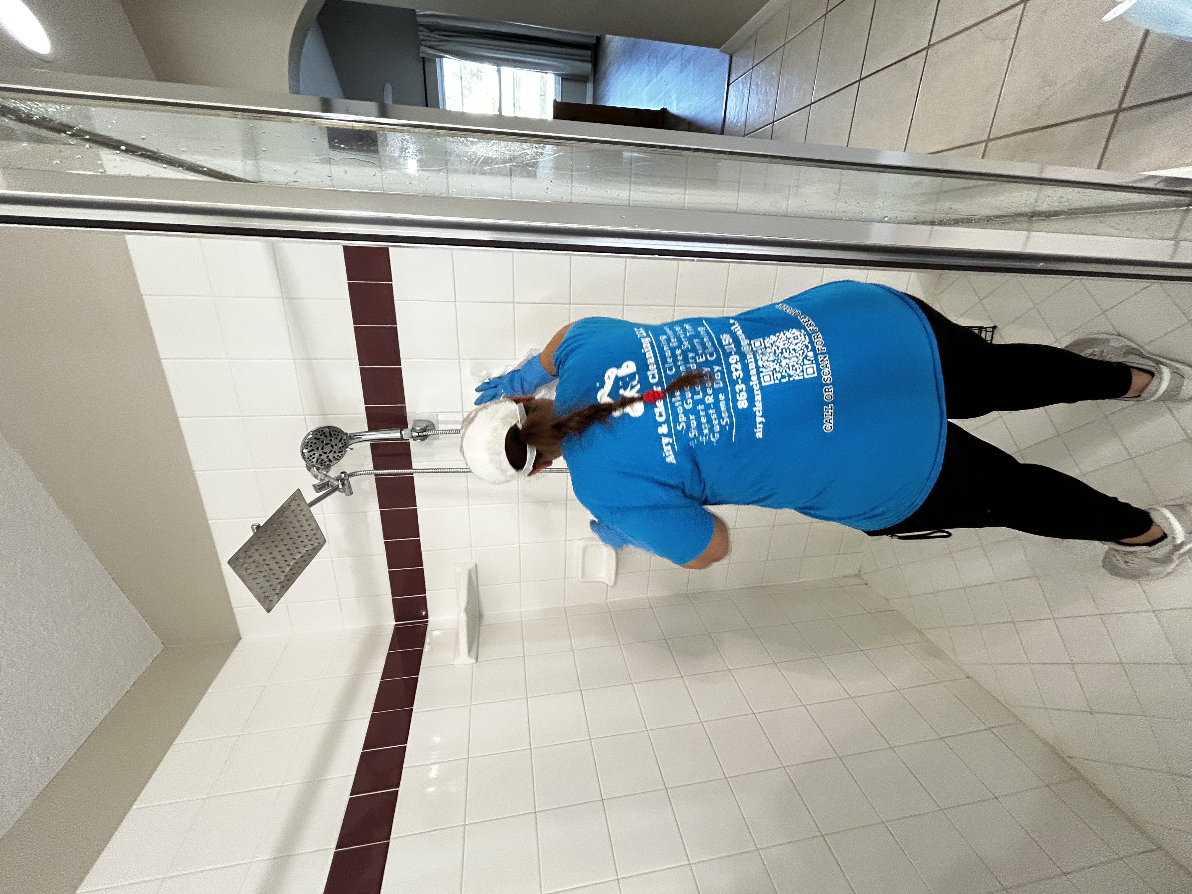 Person wearing blue shirt and gloves cleaning white tiled shower with maroon accent strip.