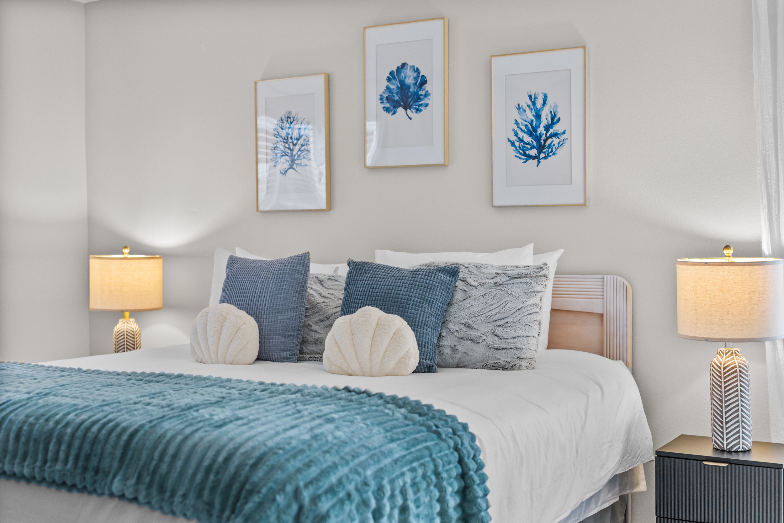 Neatly made bed with blue and white pillows, teal blanket, two bedside lamps, and three framed blue coral prints on the wall.