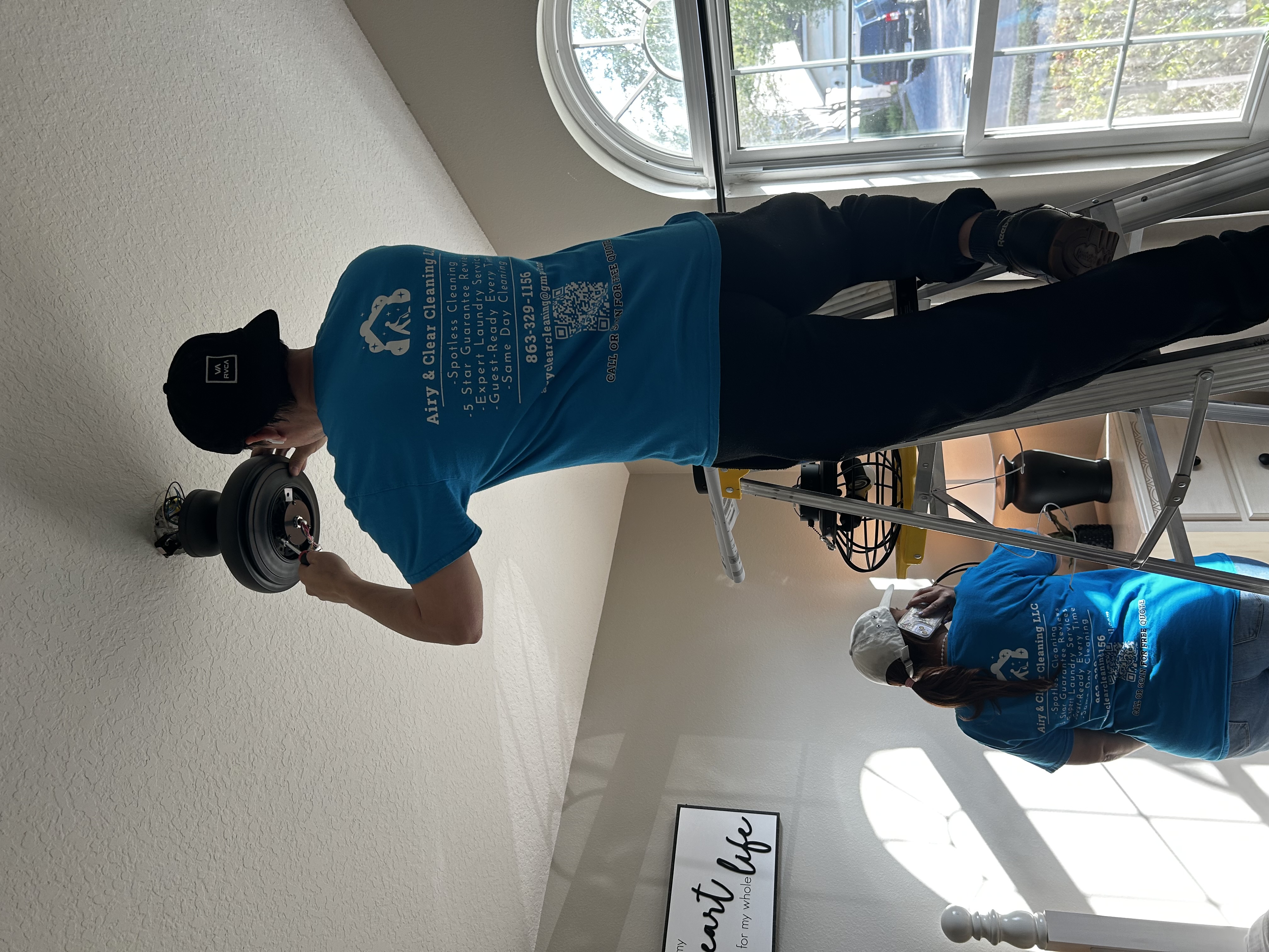 Two people in blue Airy & Clear Cleaning LLC shirts working indoors, one on a ladder fixing a ceiling fan, the other on phone.