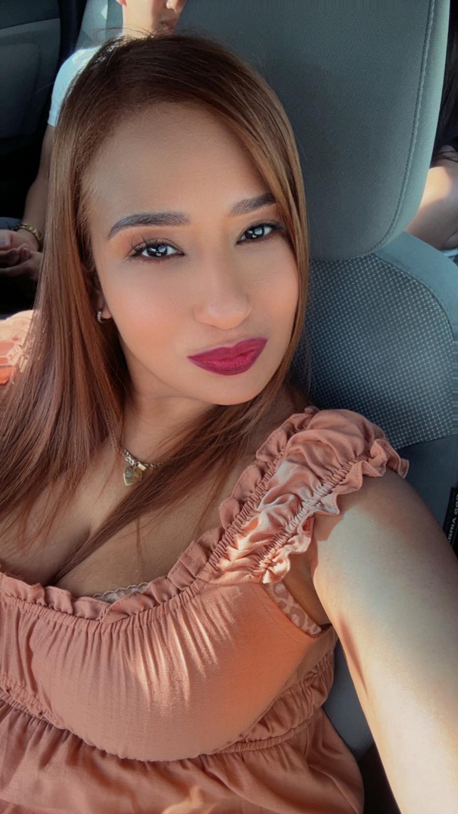 Woman with long light brown hair and bold lipstick wearing a peach ruffled top taking a selfie inside a car.