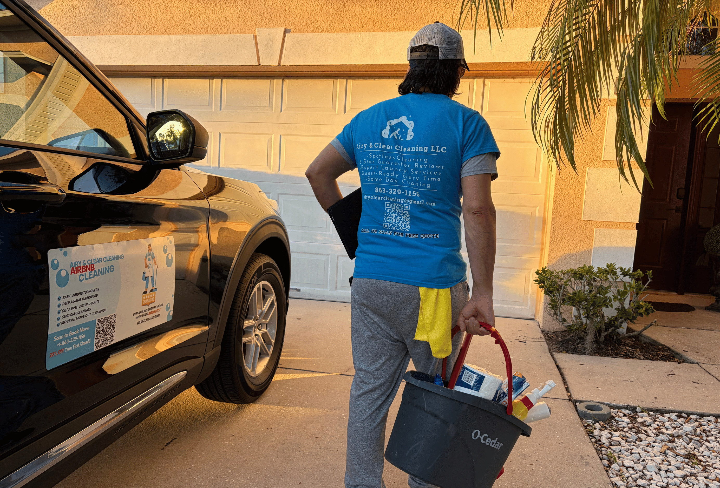Person wearing blue Airy & Clear Cleaning LLC t-shirt carrying a bucket of cleaning supplies walking towards a house with a black car parked nearby.