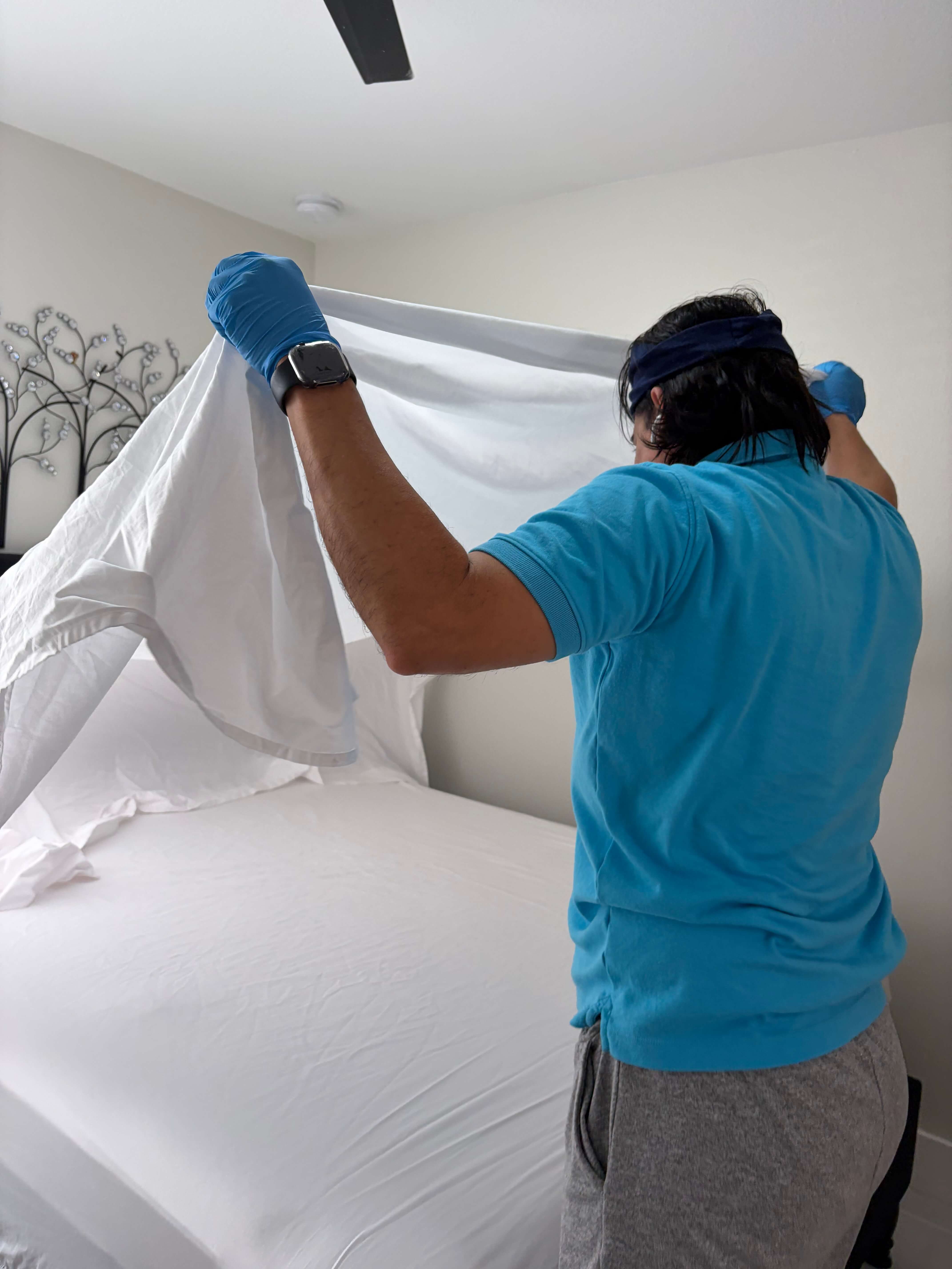 Person wearing blue gloves, a blue shirt, and gray pants making a bed with white sheets.