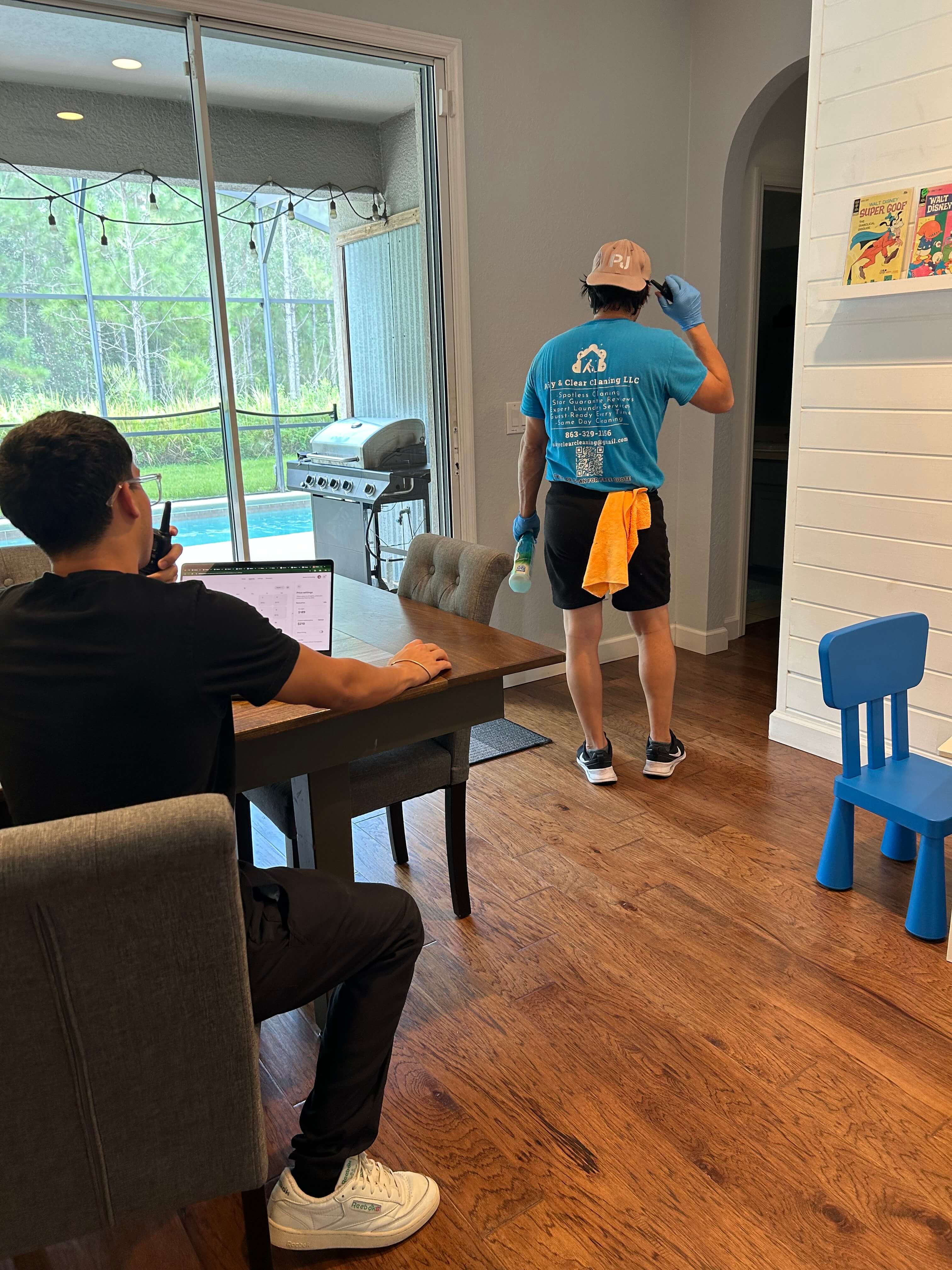 A man wearing a blue cleaning service t-shirt and gloves walks inside a house holding a spray bottle and an orange cloth, while another person sits at a dining table using a laptop and walkie-talkie.