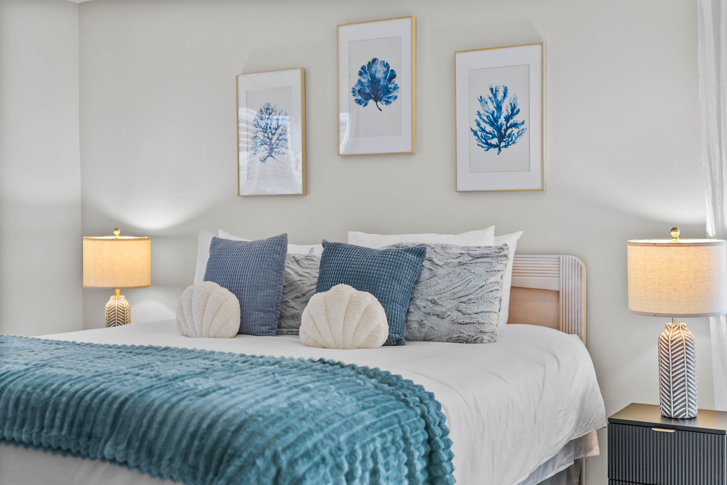 Modern bedroom with white and blue bedding, decorative pillows, two bedside lamps, and three framed blue coral prints on the wall.