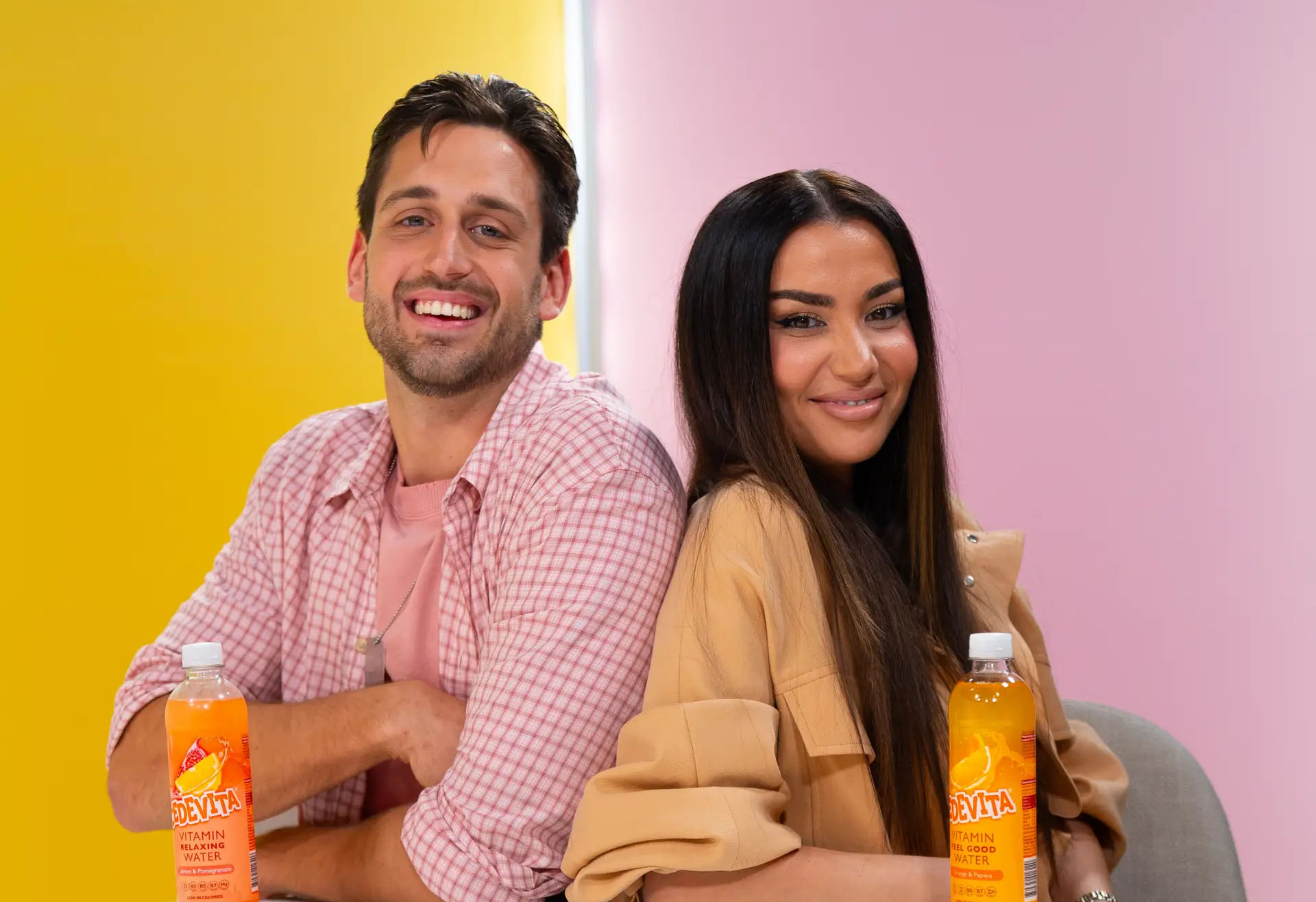 Cedevita podcast Reci tako kako je: dvoje nasmiješenih gostiju sjedi leđa uz leđa u žuto-ružičastom studiju uz boce Cedevite
