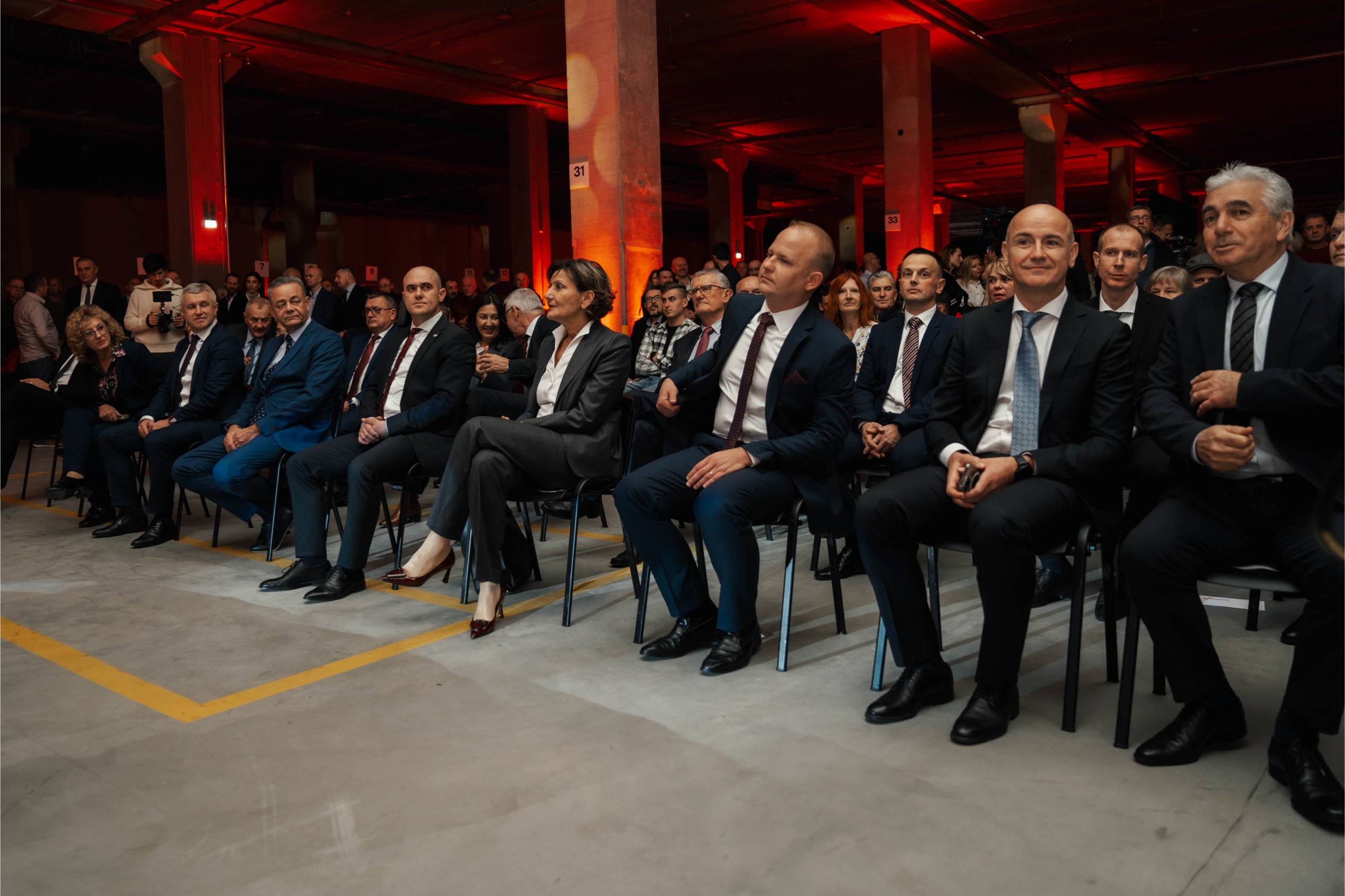 Svečano odjeveni gosti sjede u prvim redovima na ceremoniji otvorenja, iza njih narančasto osvijetljeni stupovi