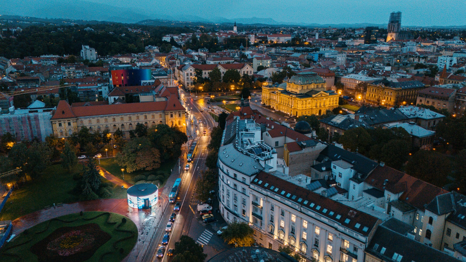 Hrvatska 2074: središte Zagreba iz zraka u sumrak, osvijetljeni okrugli paviljon uz tramvajsku prugu i HNK u pozadini