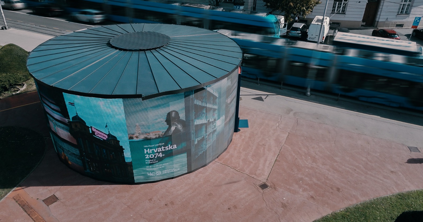 Hrvatska 2074: okrugli paviljon obložen LED ekranima iz zraka, s natpisom Hrvatska 2074. dok pokraj prolazi plavi tramvaj