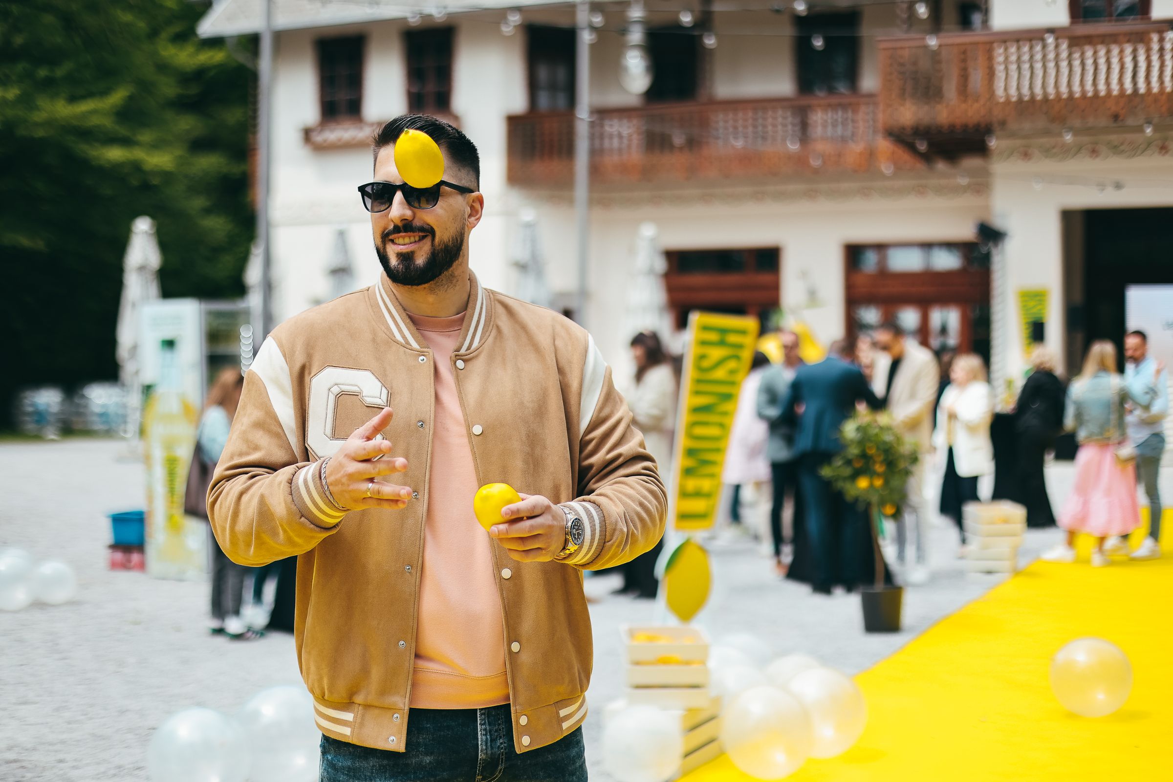 Lemonish lansiranje: muškarac sa sunčanim naočalama baca limun visoko iznad glave dok se gosti druže uz žuti tepih