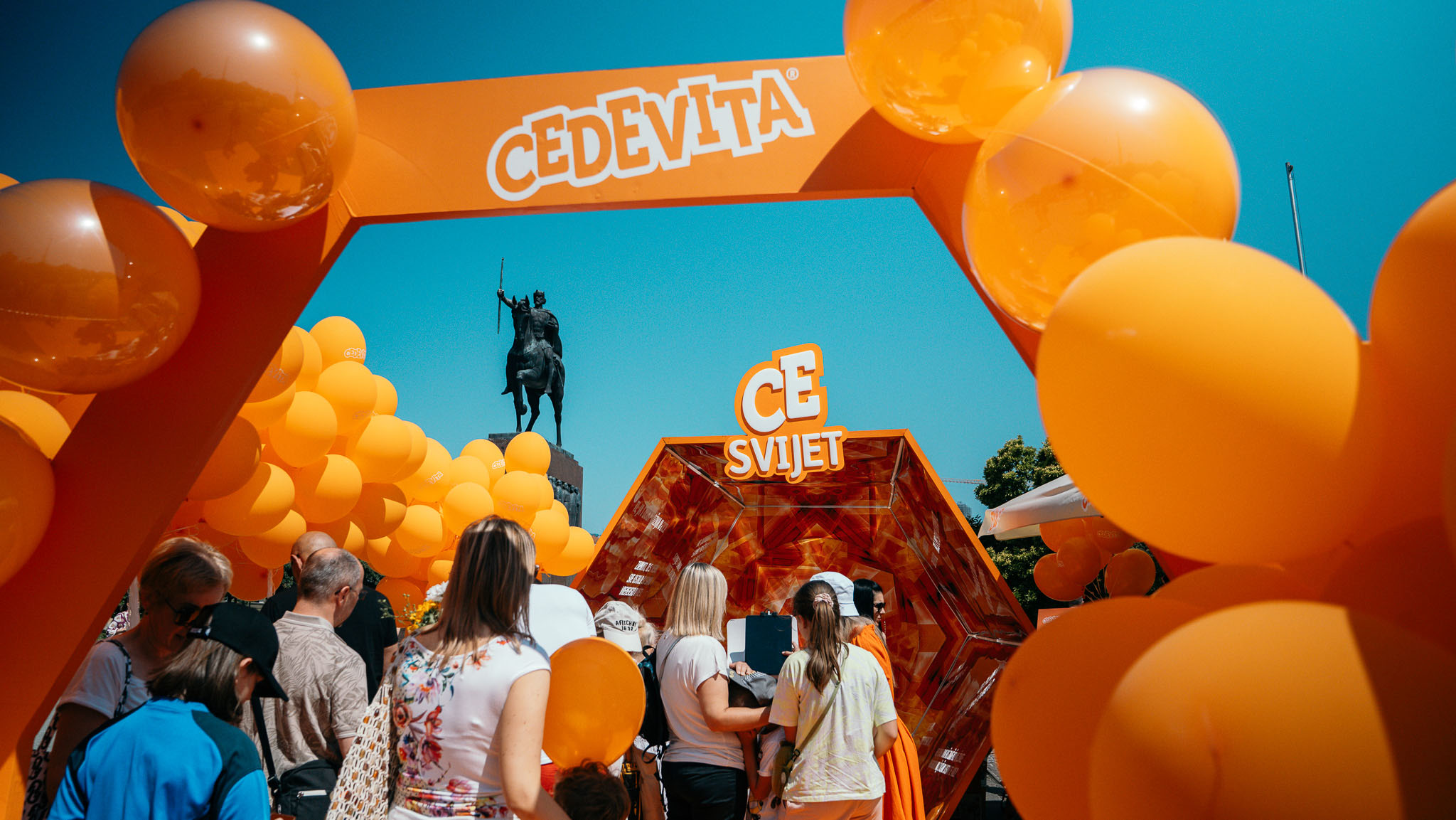 Narančasti Cedevita luk s balonima, posjetitelji okupljeni oko CE svijet kaleidoskopa, u pozadini konjanički kip