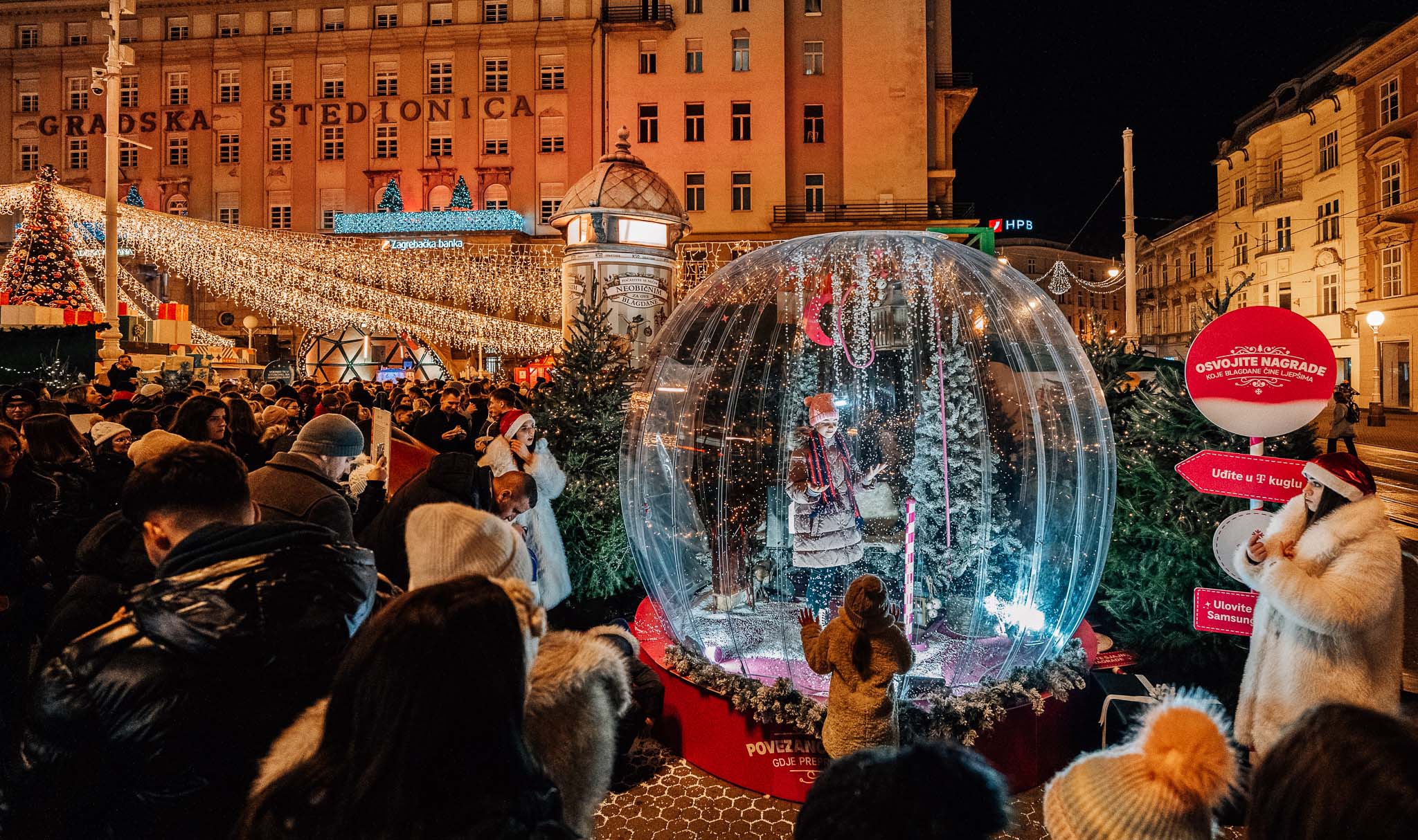 Divovska HT snježna kugla na glavnom trgu za zagrebačkog Adventa, noću, okružena posjetiteljima, lampicama i borovima