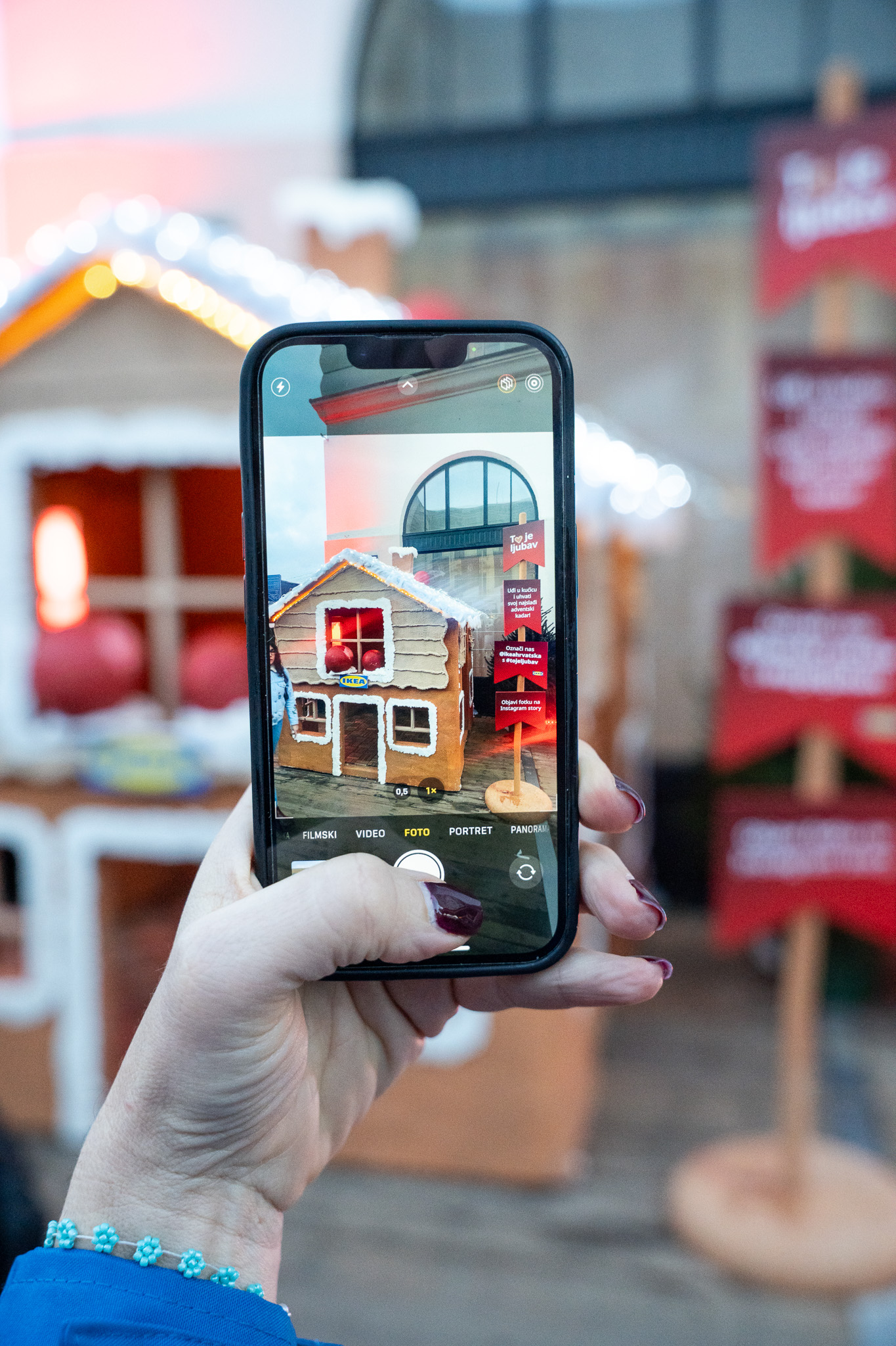 Ruka s mobitelom fotografira IKEA Vintersaga medenjak kućicu, u pozadini crveni putokazi