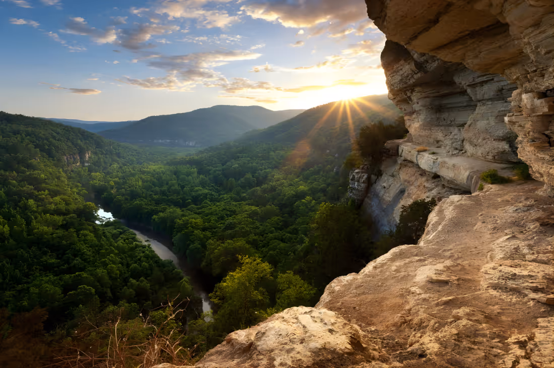 Sunset over a lush green valley with a winding river, viewed from a rocky cliff.
