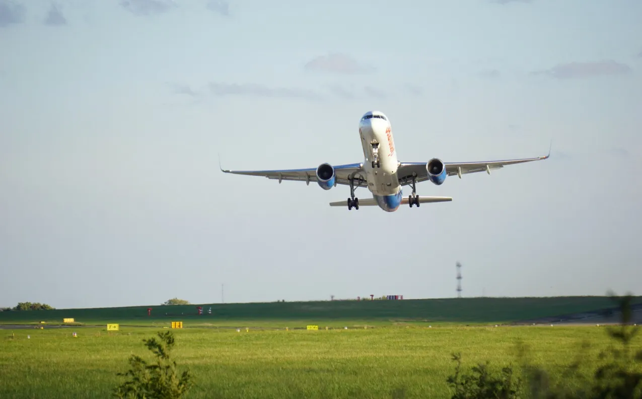 A front-facing view of a white passenger airplane with blue engine cowlings taking off from a green airfield, its nose tilted upward as it climbs into a pale blue sky.