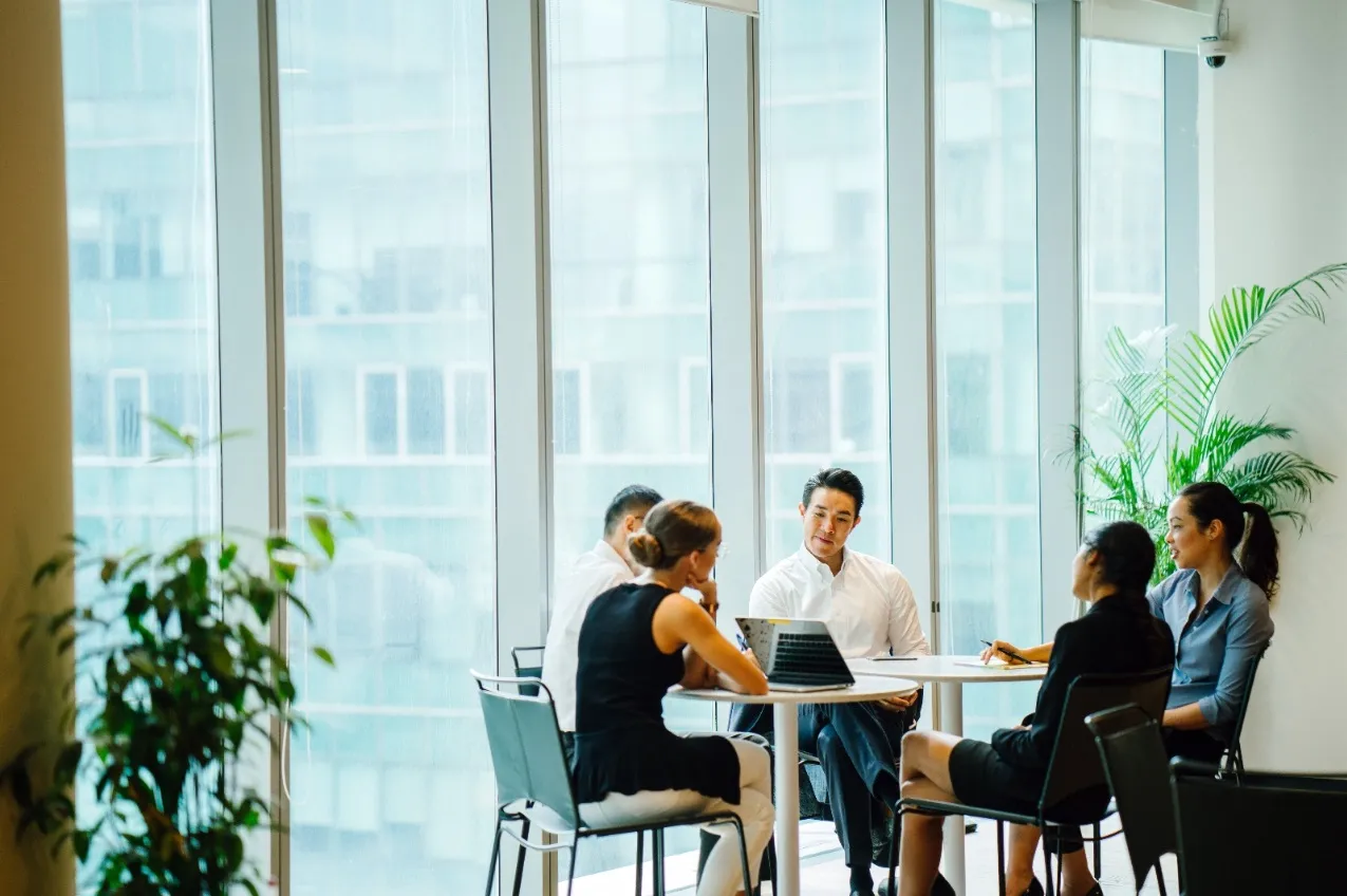 A diverse group of four business professionals in corporate attire sitting around a white circular table in a bright office. They are engaged in a meeting with a laptop open, framed by floor-to-ceiling glass windows overlooking a city.