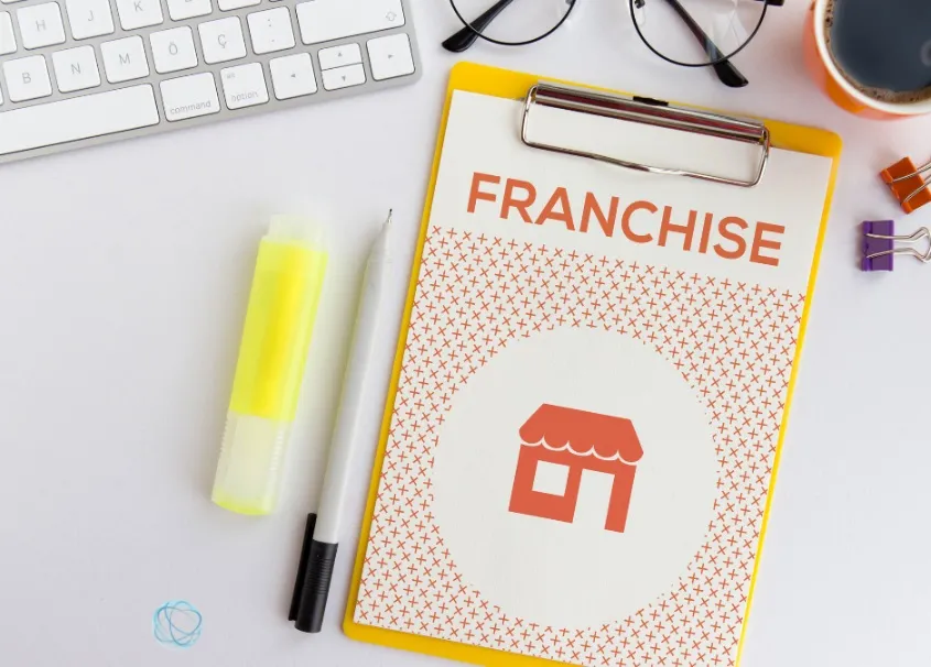 A top-down view of a white desk featuring a yellow clipboard with a paper that says 'FRANCHISE' next to a storefront icon. The desk is organized with a silver keyboard, a yellow highlighter, a pen, glasses, and a cup of coffee.