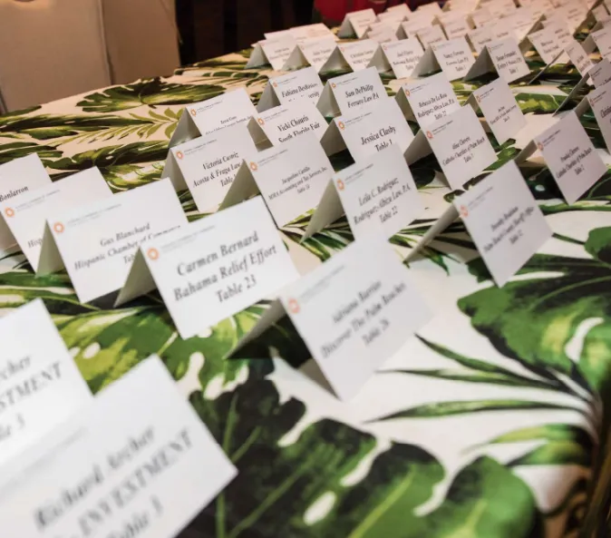 A long table covered in a tropical leaf-print tablecloth, filled with rows of white name cards for a Hispanic Chamber of Commerce networking event.