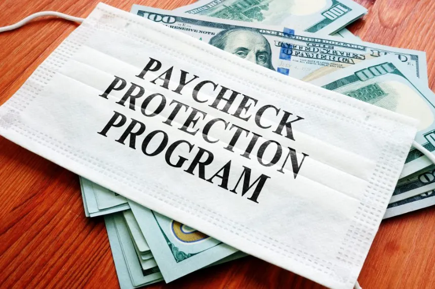 A white surgical mask with the text "PAYCHECK PROTECTION PROGRAM" resting on top of several $100 bills, illustrating the pandemic-era relief link.