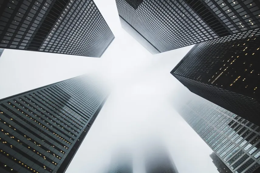 A dramatic low-angle view of towering grey and glass skyscrapers reaching into a misty white sky, evoking a professional corporate environment.