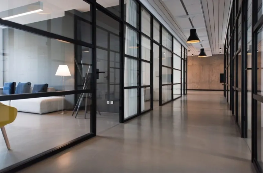 A sleek, modern office hallway with polished concrete floors, glass-walled meeting rooms with black frames, and minimalist pendant lighting.