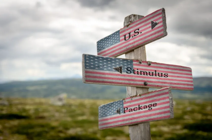 A wooden signpost in a grassy field with three directional arrows painted as American flags with the words "U.S.," "Stimulus," and "Package."