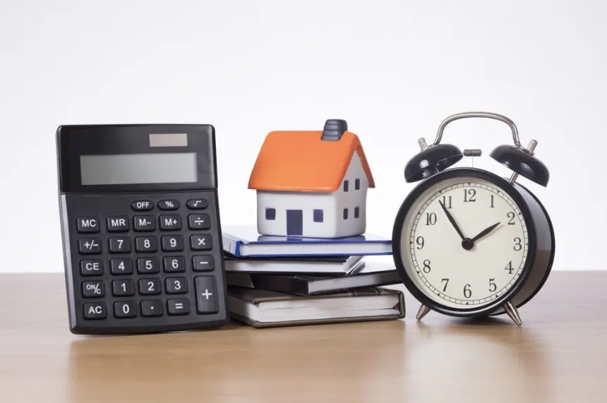 A black office calculator, a small model house with an orange roof, and a black alarm clock arranged together on a wooden desk.