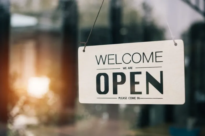A wooden "WELCOME WE ARE OPEN" sign hanging on a glass door, with a warm bokeh light effect in the background.