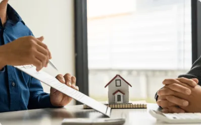 A close-up of an attorney in a suit sitting across from a client, using a pen to guide them through a real estate contract next to a small model house on the desk.