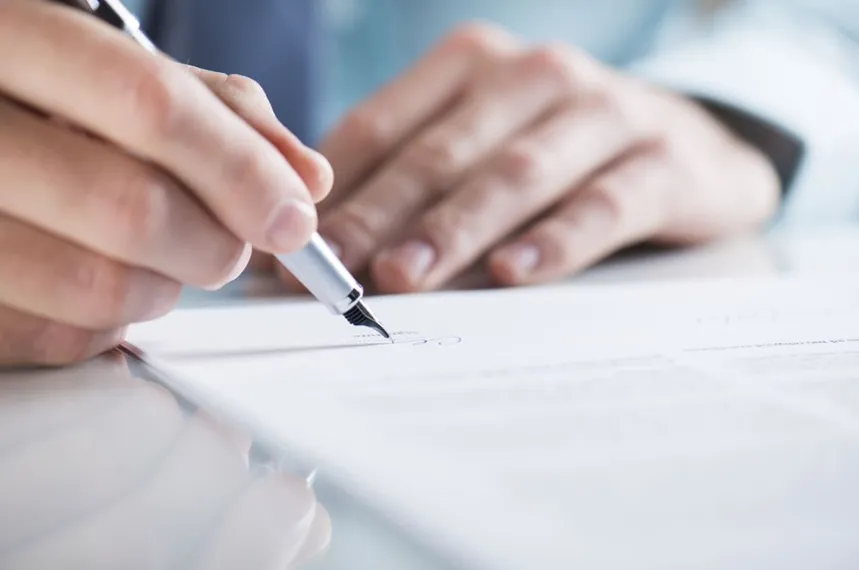 A close-up of a person's hand using a silver fountain pen to sign a formal legal document on a bright white desk.