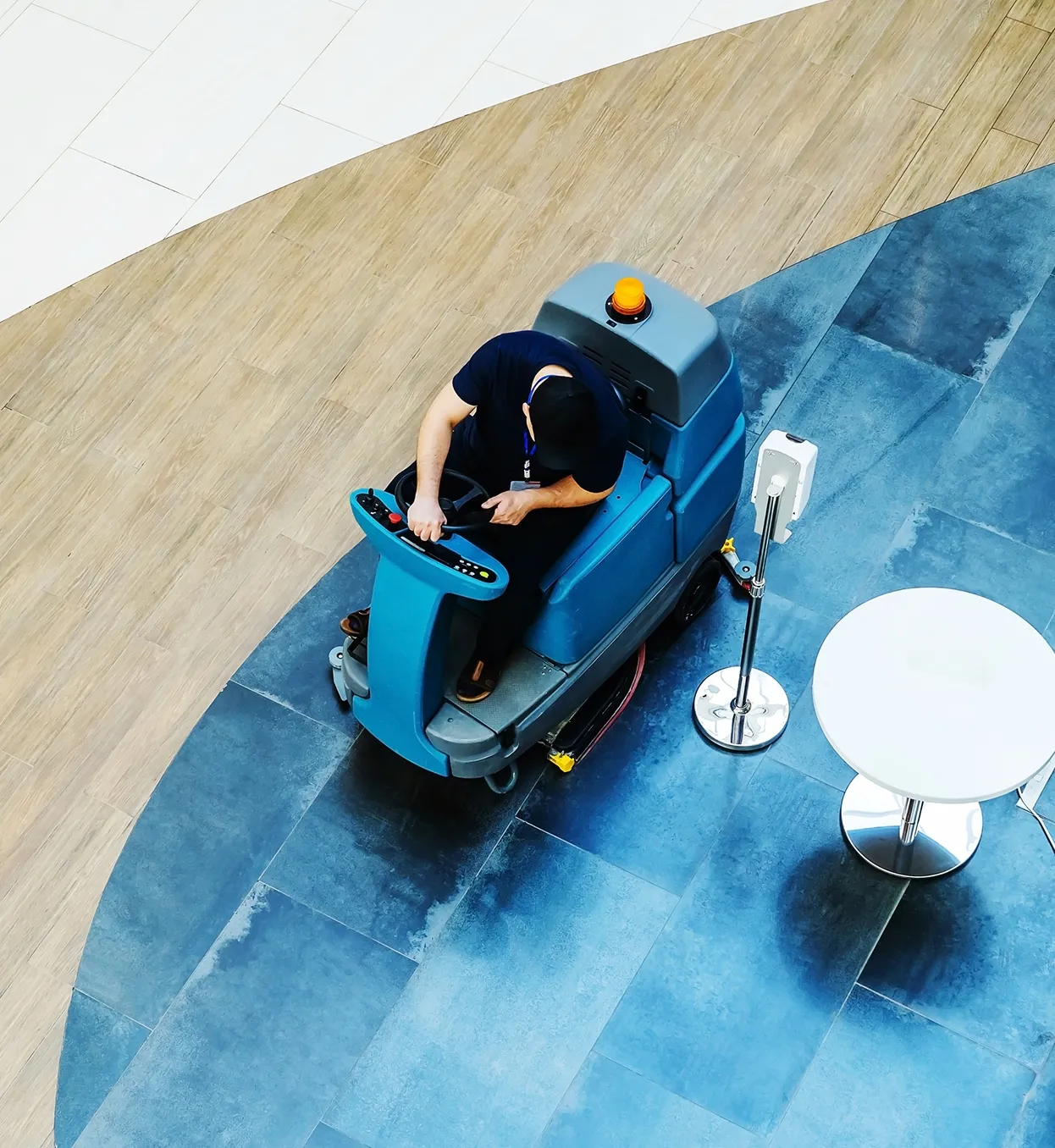 Person operating a blue floor cleaning machine on dark blue and light wood patterned flooring near a white round table and a hand sanitizer stand.