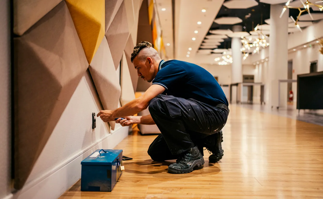 Man kneeling on a wooden floor working on an electrical outlet with a screwdriver beside a blue toolbox in a modern room with geometric wall panels.