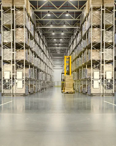 Interior of a large warehouse with tall metal shelves stocked with pallets and a yellow forklift lifting a pallet in the center aisle.