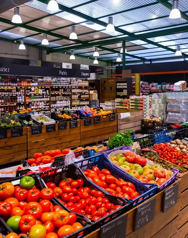 Indoor grocery store produce section with wooden crates filled with various tomatoes, leafy greens, and other vegetables under bright ceiling lights.