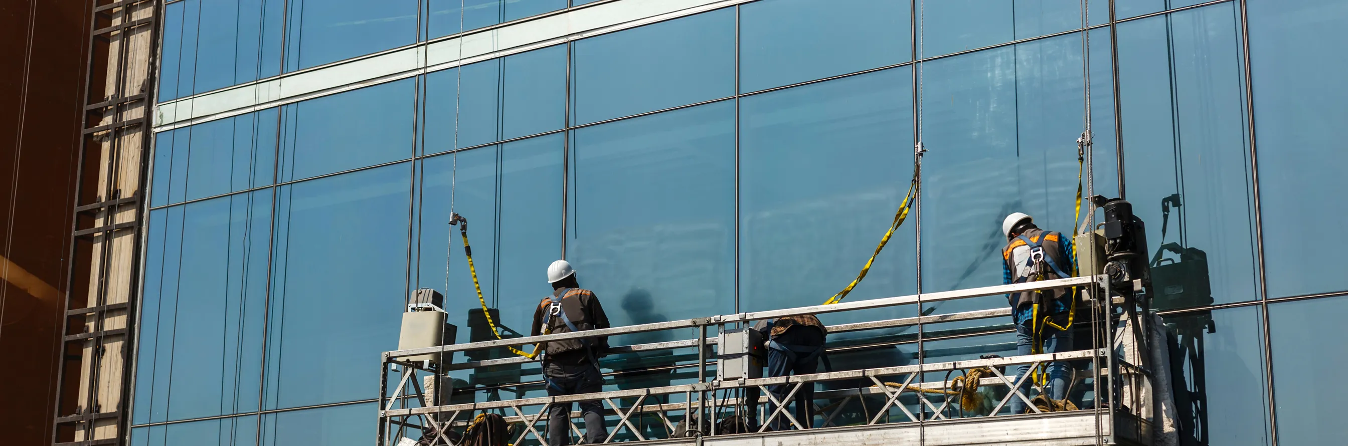 Two workers wearing safety gear on a suspended platform cleaning a glass building facade.