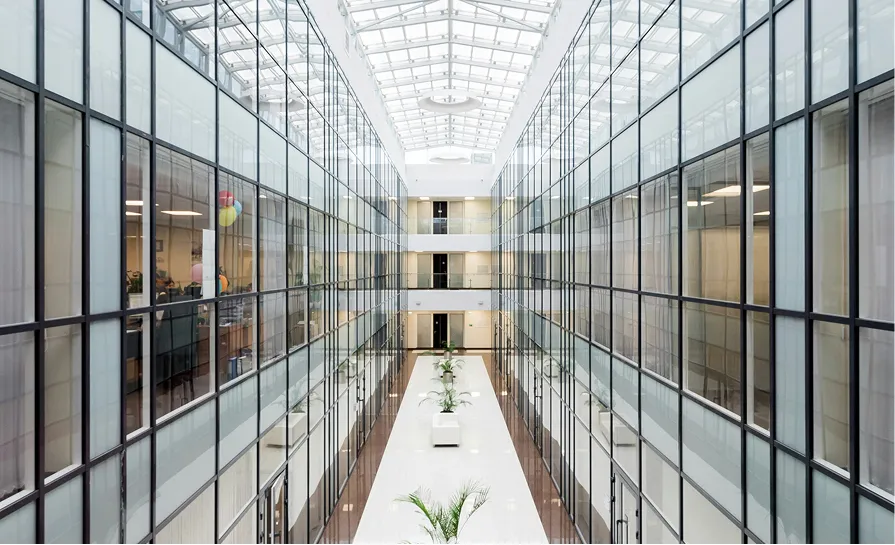 Bright modern office atrium with glass walls, white floors, indoor plants, and a glass ceiling.